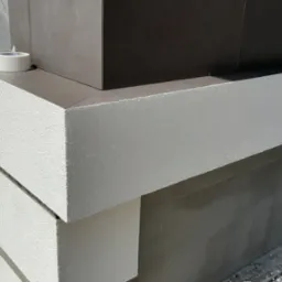 Wykończony fragment elewacji budynku z szarymi płytkami i białym tynkiem strukturalnym, na rogu leży rolka taśmy malarskiej.