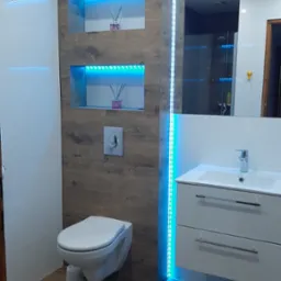 Nowoczesna łazienka z drewnopodobną ścianą, podświetlanymi wnękami i lustrem, wisząca toaleta i biała szafka z umywalką.