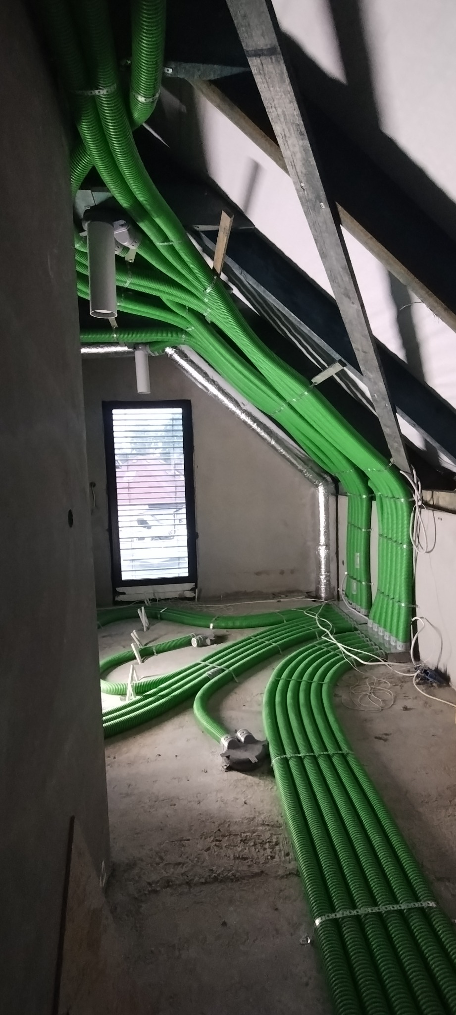 Wnętrze poddasza w trakcie instalacji systemu wentylacyjnego z licznymi zielonymi, karbowanymi przewodami wentylacyjnymi, częściowo przymocowanymi do sufitu i ścian, z widocznym oknem z żaluzjami...