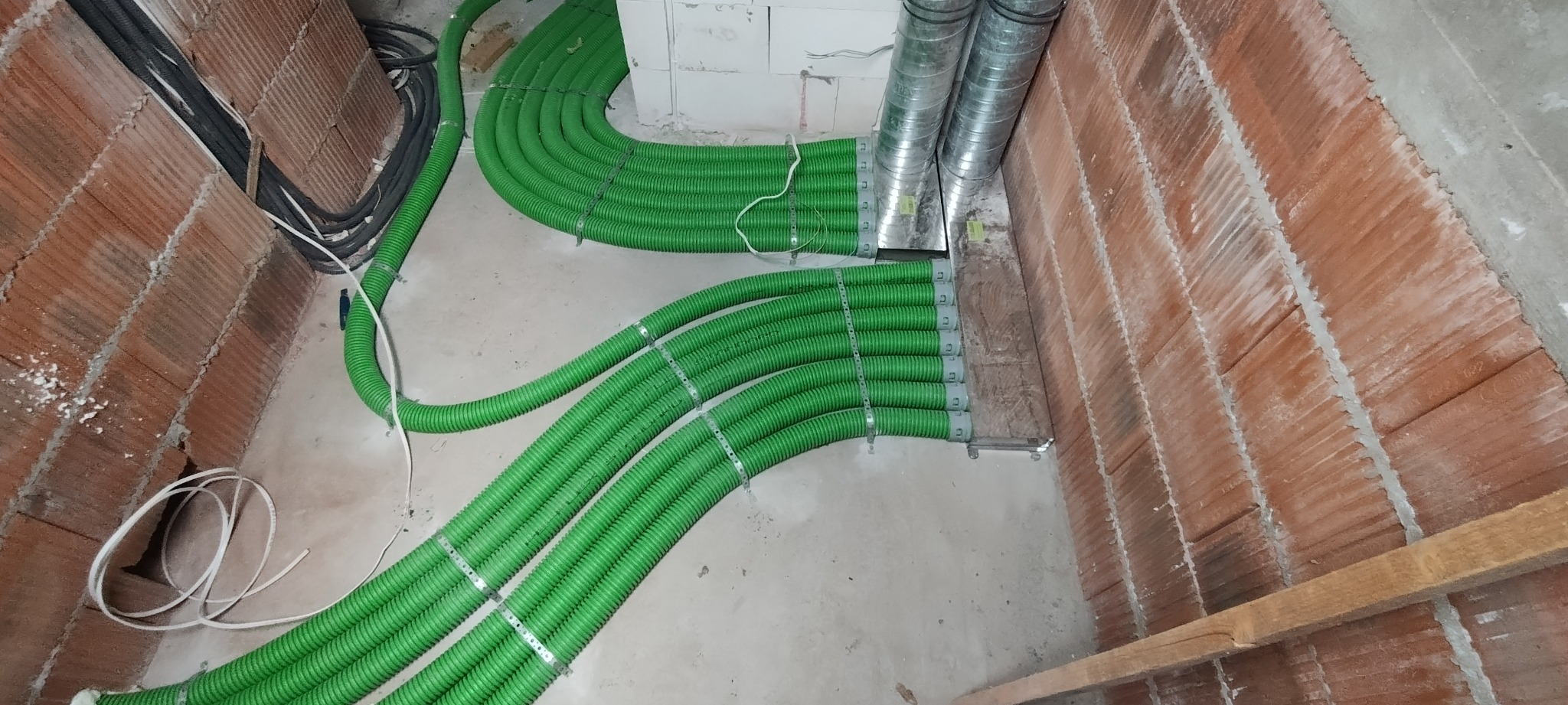 Zielone, karbowane rury wentylacyjne przymocowane metalowymi obejmami, ułożone równolegle w surowym wnętrzu z ceglanymi ścianami i betonową podłogą, w tle widoczne czarne kable.