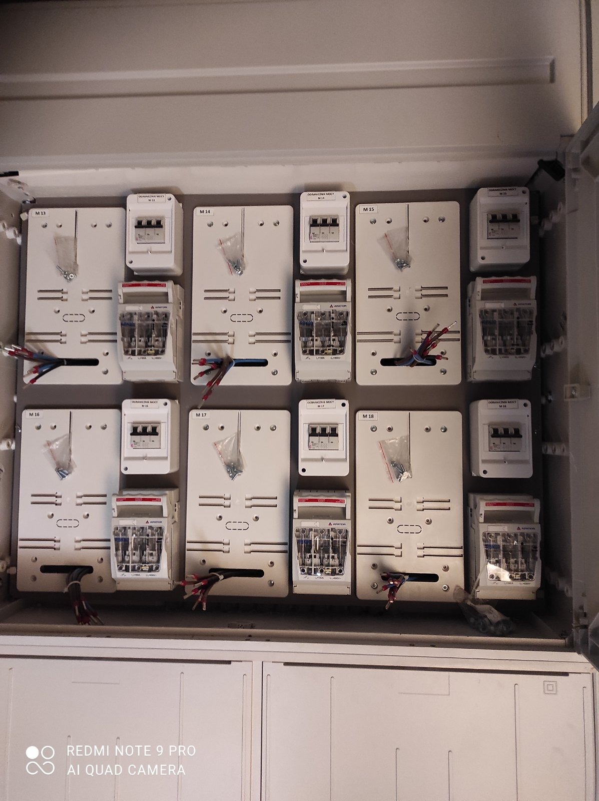 Rozdzielnica elektryczna w trakcie montażu, widoczne zabezpieczenia nadprądowe, ograniczniki mocy i przewody w skrzynce z numeracją M13 do M18, niektóre z woreczkami ze śrubkami.