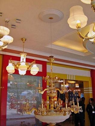 Wielopoziomowy, bogato zdobiony żyrandol z elementami złota, wiszący w pomieszczeniu z czerwono-żółtymi ścianami i innymi lampami w tle.