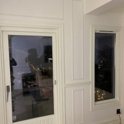 DEFO Kacper Dębczak - Białe, wykończone listwami ściany z drzwiami balkonowymi i oknem, widoczne odbicia w szybach, na podłodze wiadro z farbą i kable.