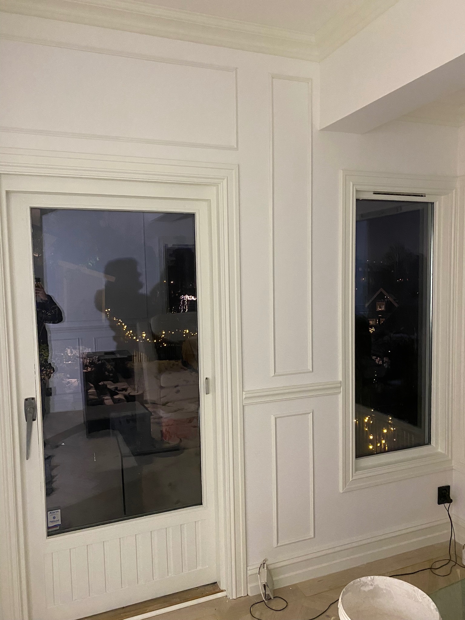 Białe, wykończone listwami ściany z drzwiami balkonowymi i oknem, widoczne odbicia w szybach, na podłodze wiadro z farbą i kable.