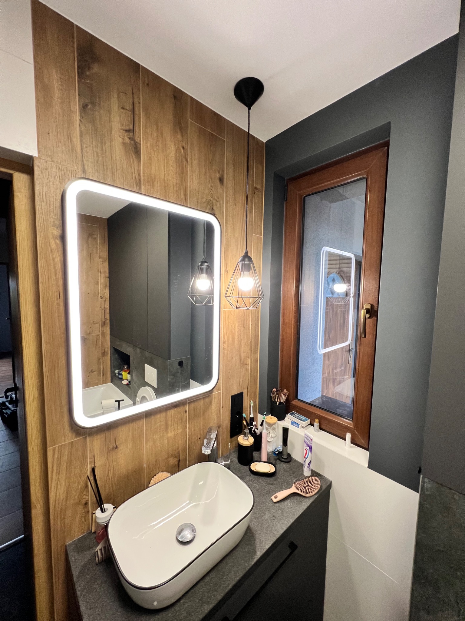 Nowoczesna łazienka z drewnopodobnymi płytkami na ścianie, owalną umywalką nablatową, podświetlanym lustrem i wiszącą lampą drucianą.