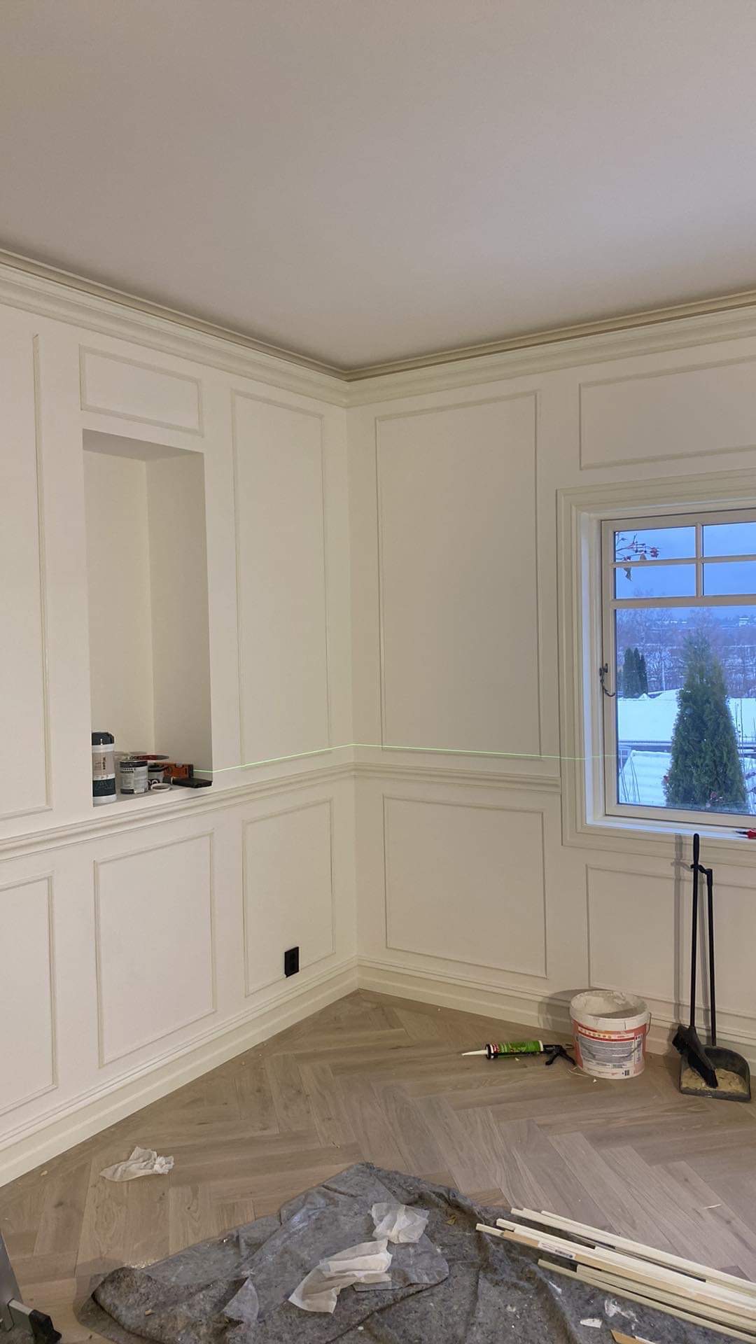 Pokój z panelami ściennymi w stylu klasycznym, podłoga w jodełkę, widoczny laser krzyżowy, puszka farby, tuba kleju i listwy przypodłogowe przygotowane do montażu.