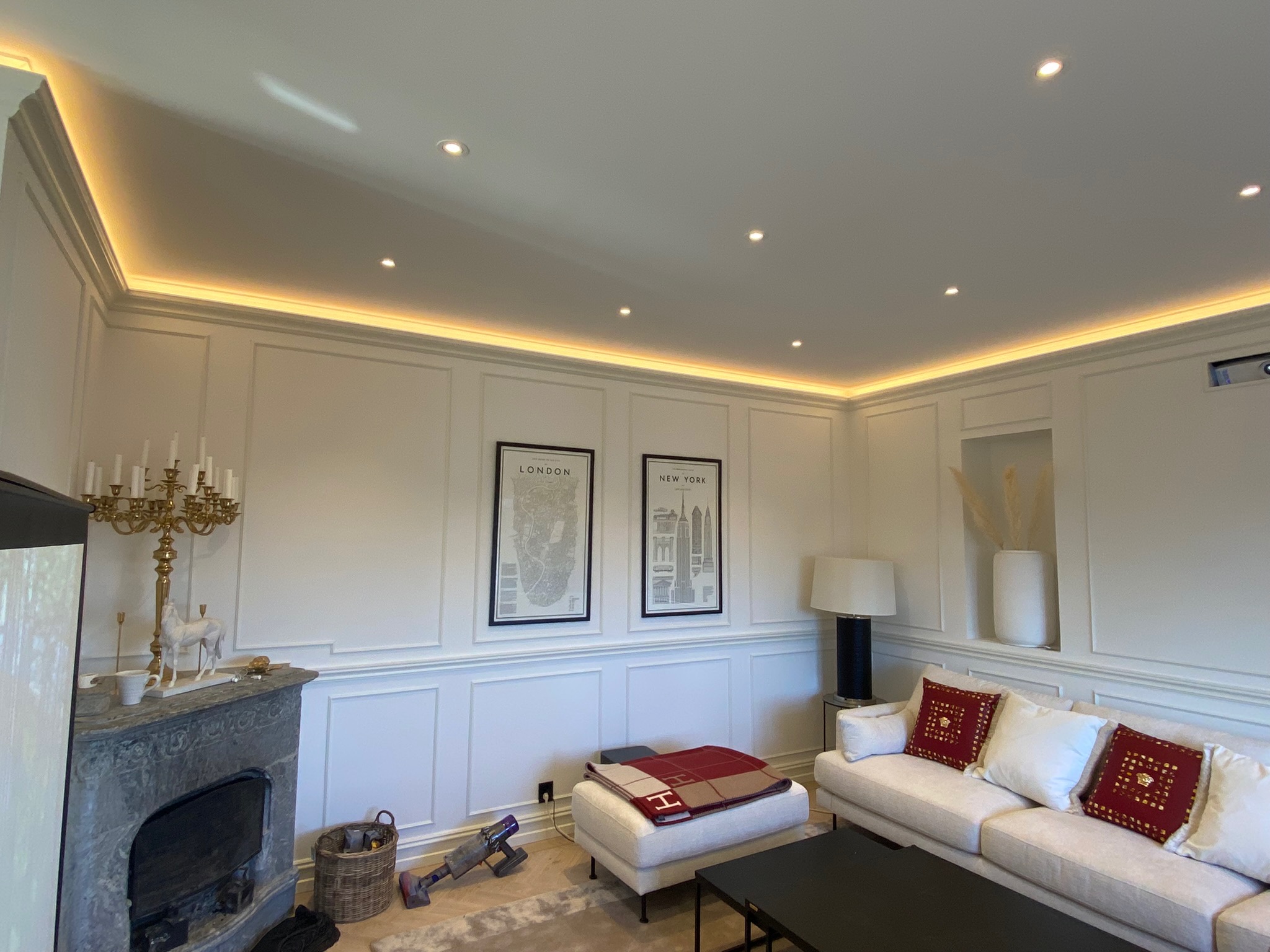 Elegancki salon z dekoracyjną sztukaterią ścienną, oświetleniem LED podsufitowym, marmurowym kominkiem, sofą z ozdobnymi poduszkami i plakatami w ramkach. Widoczny odkurzacz akumulatorowy.