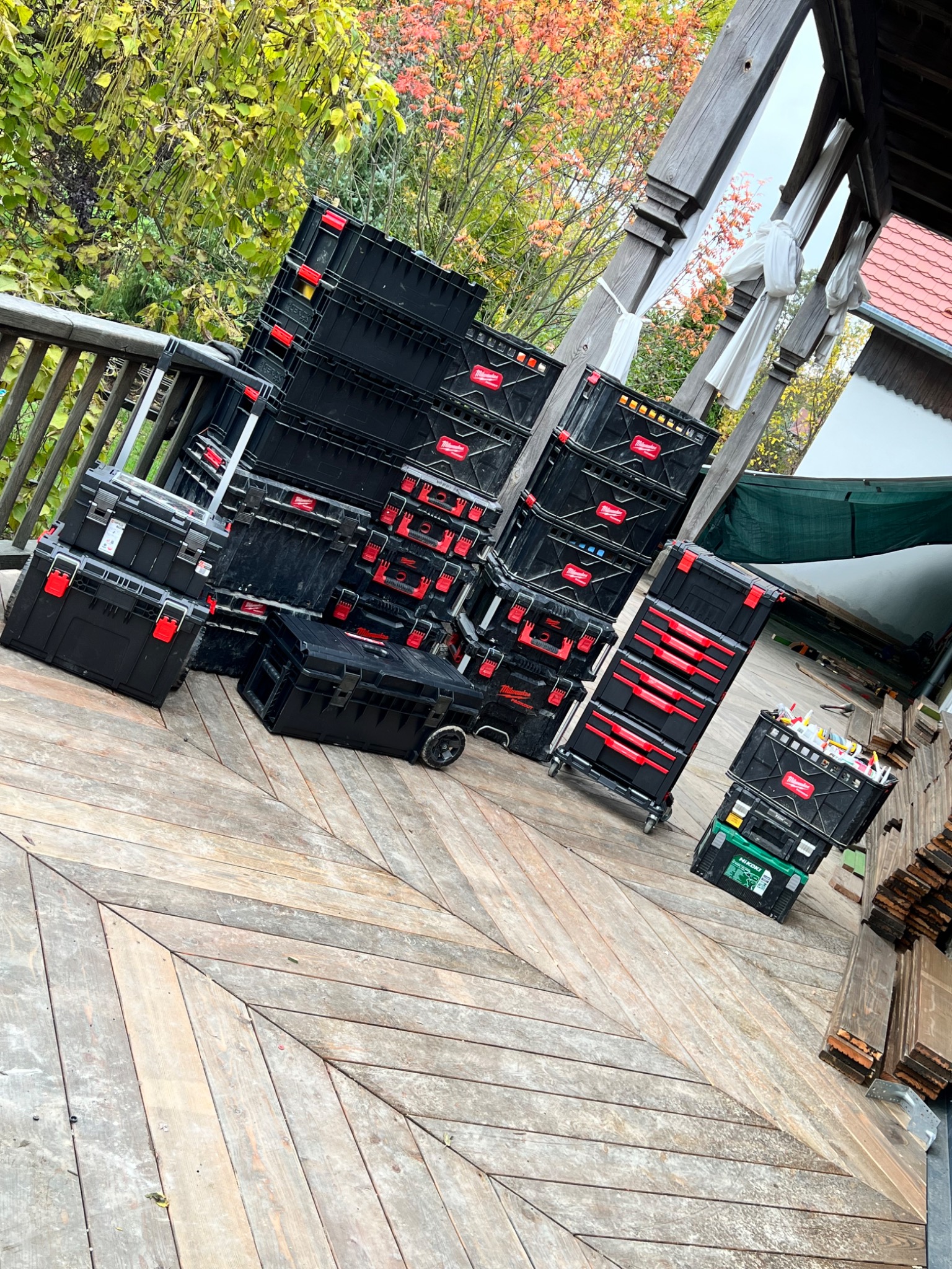Stos czarnych skrzynek narzędziowych Milwaukee z czerwonymi akcentami na drewnianym tarasie z widokiem na jesienne drzewa.