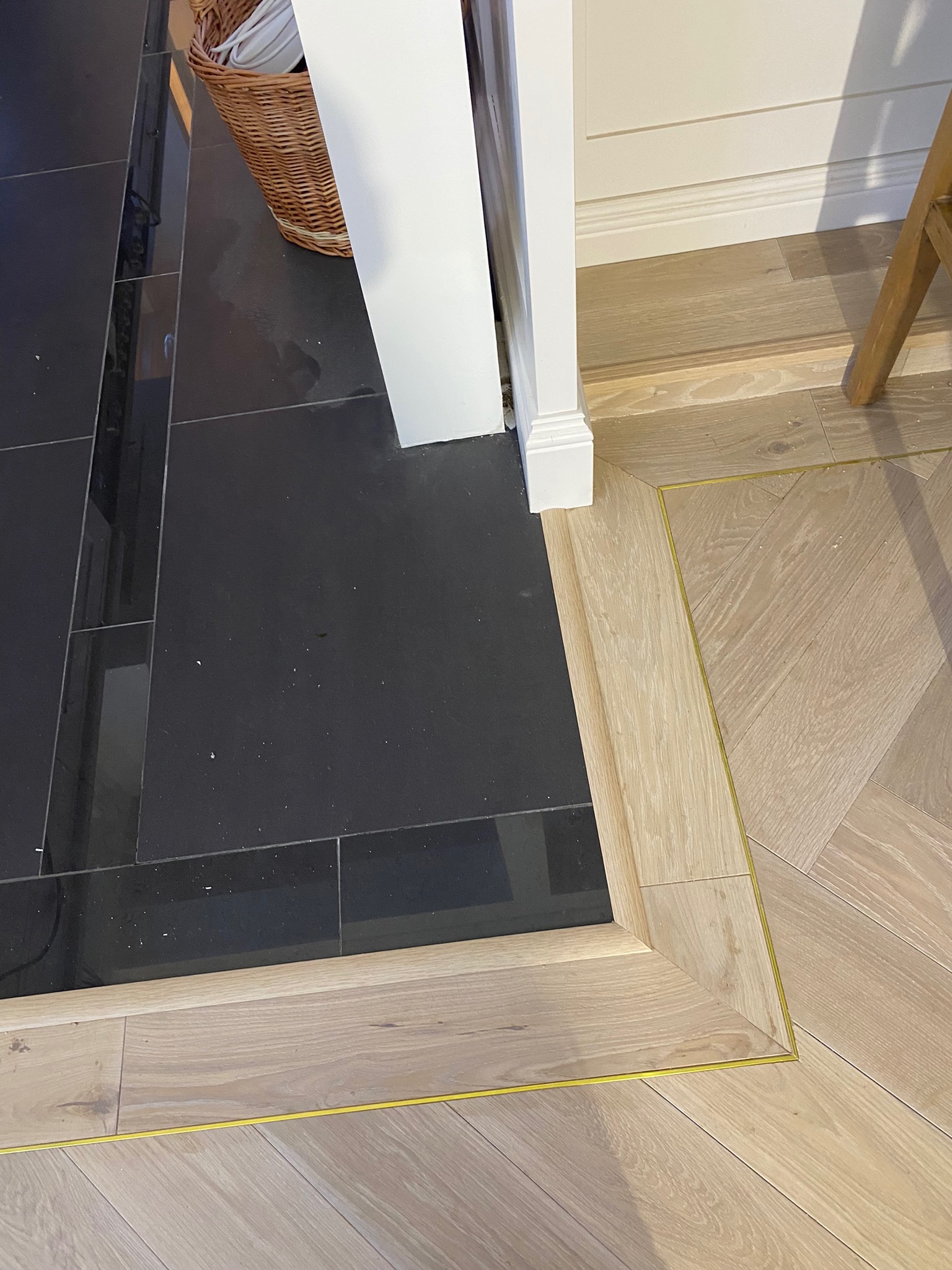 Kontrastowe połączenie czarnej, błyszczącej terakoty i jasnej, drewnianej podłogi ułożonej we wzór jodełki, oddzielone listwą z delikatnym złotym akcentem przy framudze drzwi.