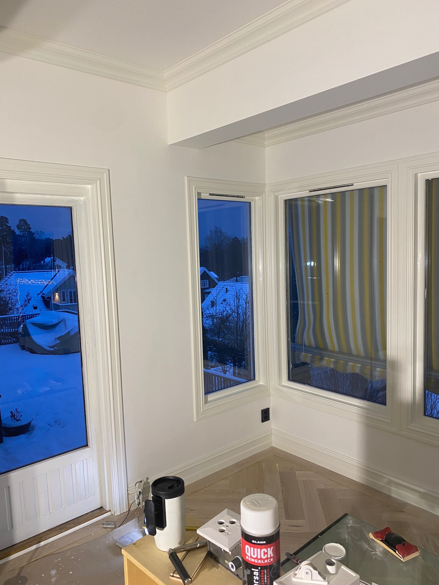 Wnętrze pokoju po malowaniu ścian i listew okiennych na biało, widoczne narzędzia malarskie na stoliku, parkiet w jodełkę na podłodze, za oknem zimowy krajobraz.