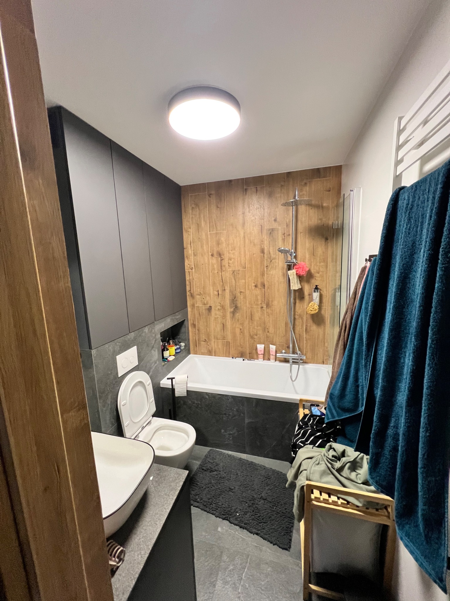 Nowoczesna łazienka z drewnianymi akcentami: płytki imitujące drewno na ścianie przy wannie, szare szafki wiszące, biała umywalka nablatowa, toaleta i grzejnik drabinkowy z wiszącymi ręcznikami.