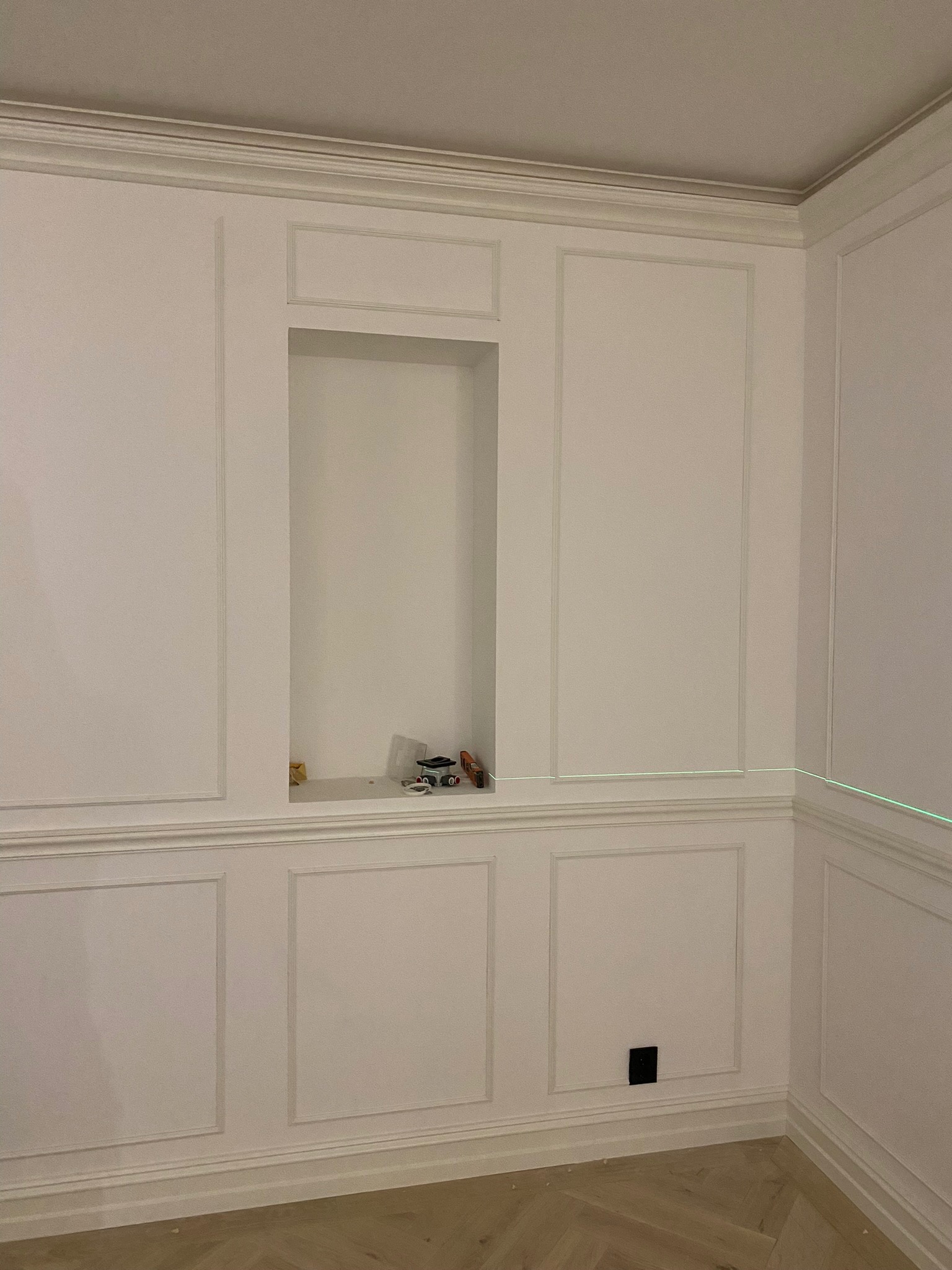 Ściana z dekoracyjnymi panelami w stylu angielskim, z wnęką i widoczną linią lasera poziomującego, w trakcie remontu.