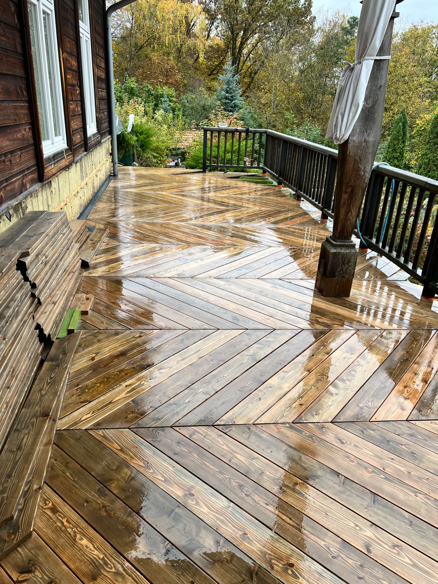 Świeżo zaimpregnowany drewniany taras z wzorem jodełki, częściowo mokry od deszczu, z balustradą i słupem podtrzymującym, widoczne deski do dalszej budowy.