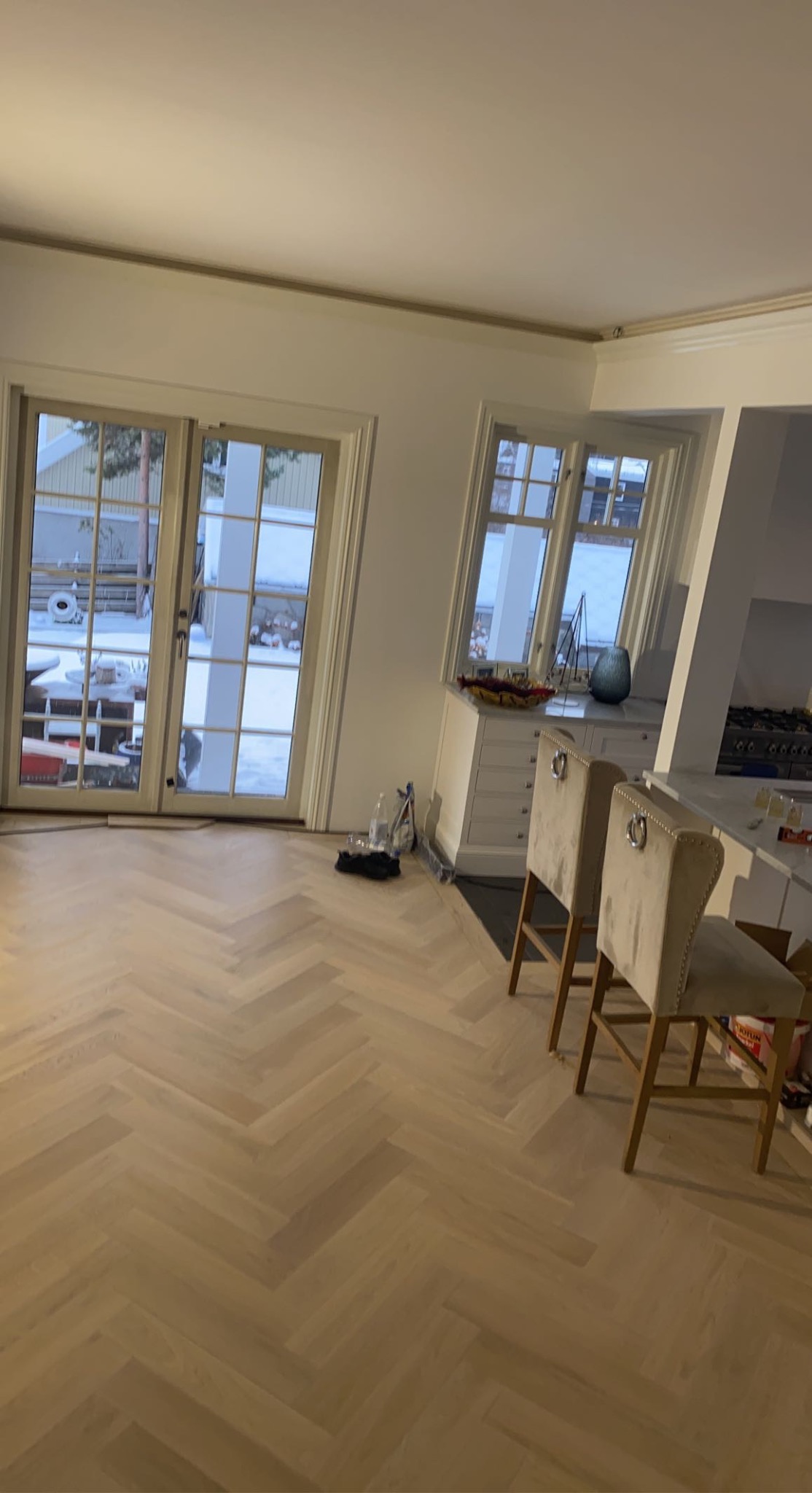 Jasna, nowo ułożona podłoga z paneli ułożonych we wzór jodełki francuskiej w przestronnym wnętrzu, widoczne drzwi balkonowe i krzesła barowe