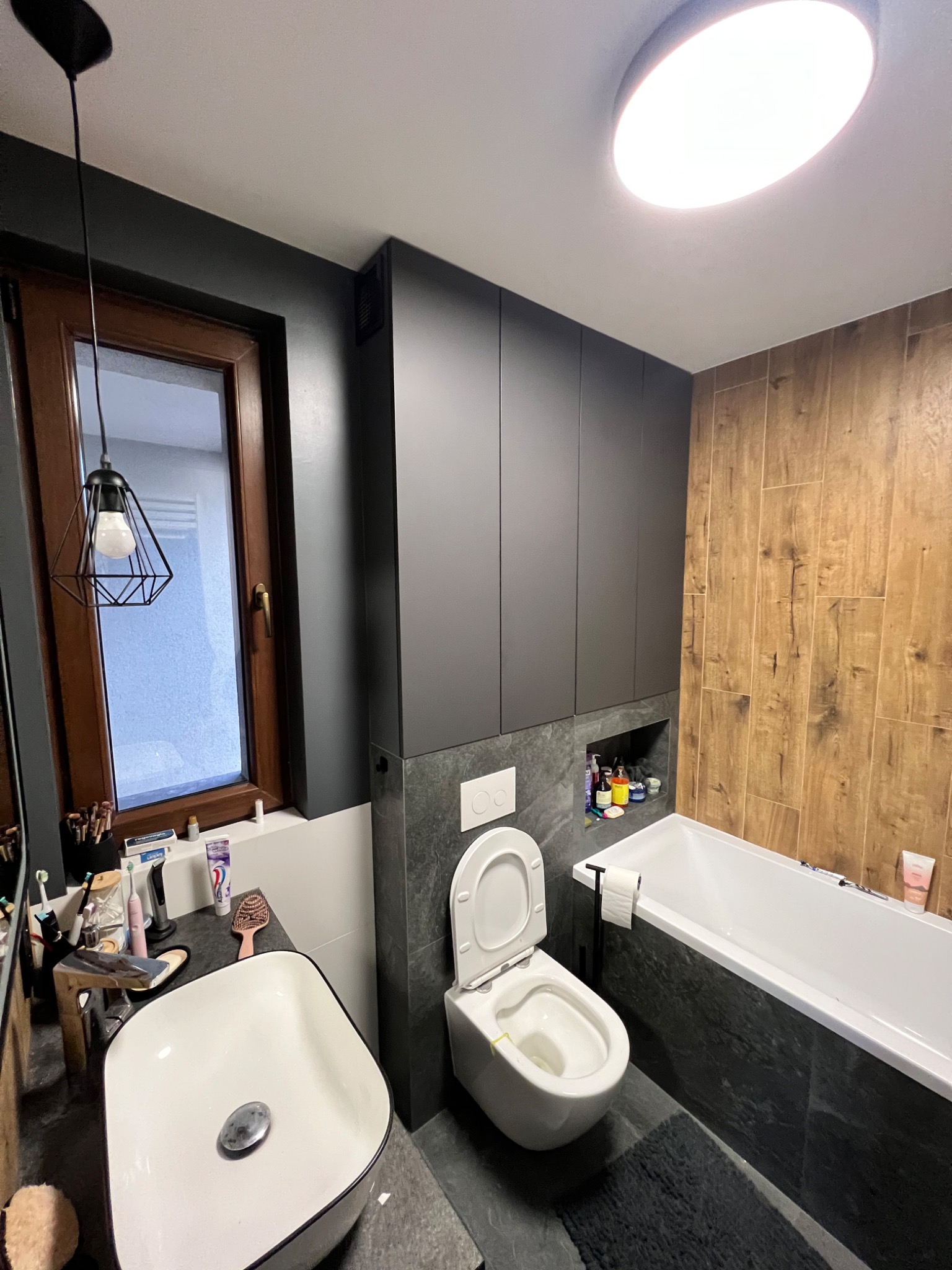 Nowoczesna łazienka z drewnopodobnymi płytkami, zabudową szafek w kolorze grafitowym, wiszącą toaletą i umywalką nablatową.