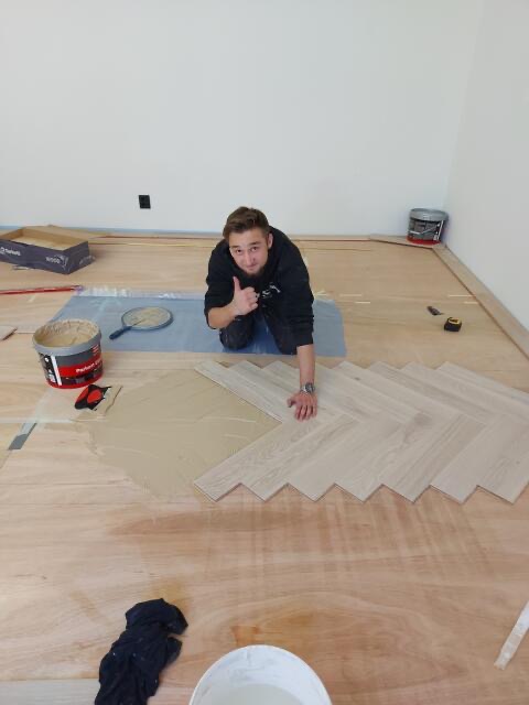 Mężczyzna w trakcie układania parkietu w jodełkę, kładzie klej i układa deski, kciuk w górę, widoczne narzędzia i materiały.