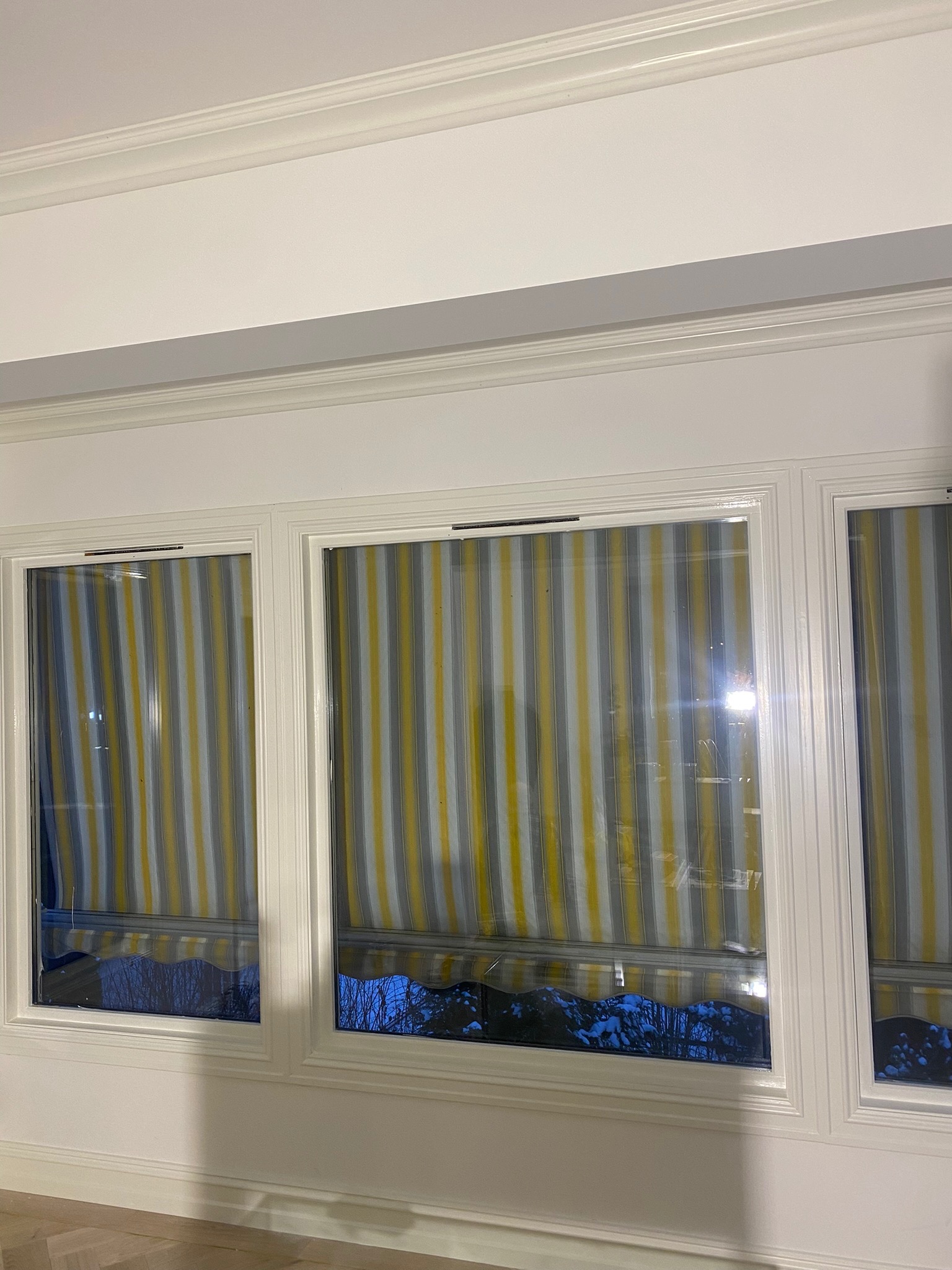 Trzy okna z zamontowanymi roletami w biało-szaro-żółte pionowe pasy, widok na zaśnieżony ogród, białe ramy okienne z dekoracyjnymi listwami.