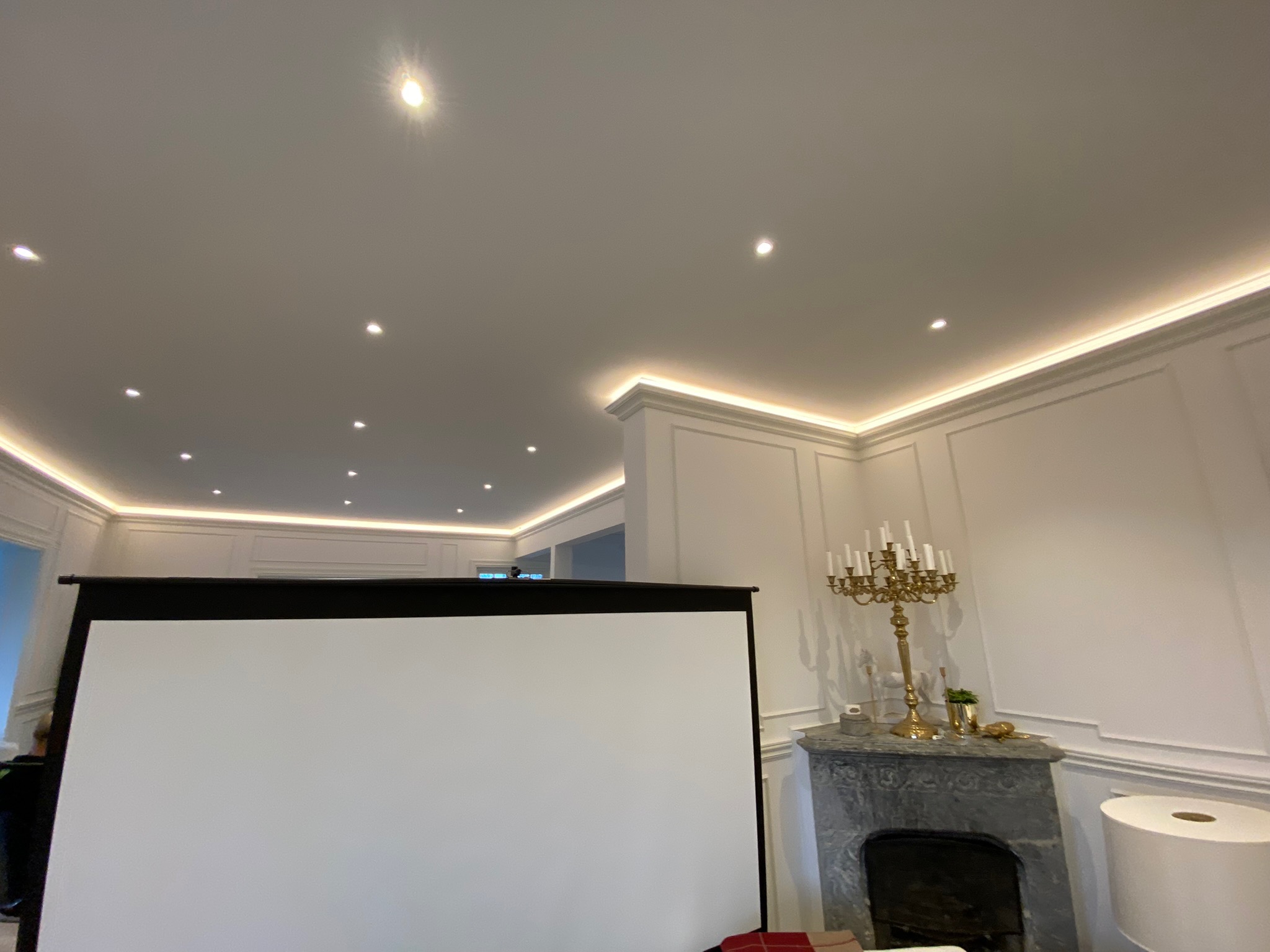 Eleganckie wnętrze z oświetleniem LED podsufitowym, sztukaterią ścienną i marmurowym kominkiem z dekoracyjnym świecznikiem, zasłonięte częściowo przez rozłożony ekran projekcyjny.