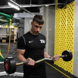 Młody mężczyzna w czarnej koszulce z krótkim rękawem, ćwiczący biceps sztangą łamaną w siłowni, z widocznymi mięśniami ramion i sprzętem do ćwiczeń w tle.