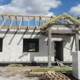 Surowy stan budynku z betonu komórkowego z widoczną konstrukcją więźby dachowej z drewnianych belek, palety z bloczkami Prefabet na pierwszym planie, okna i drzwi bez stolarki.