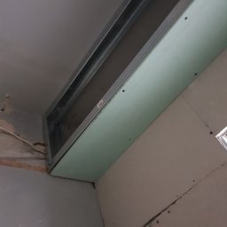 Adam - Montaż płyty kartonowo-gipsowej na metalowej konstrukcji sufitowej w trakcie remontu, widoczne śruby mocujące i fragment starej ściany w tle.