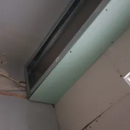 Montaż płyty kartonowo-gipsowej na metalowej konstrukcji sufitowej w trakcie remontu, widoczne śruby mocujące i fragment starej ściany w tle.