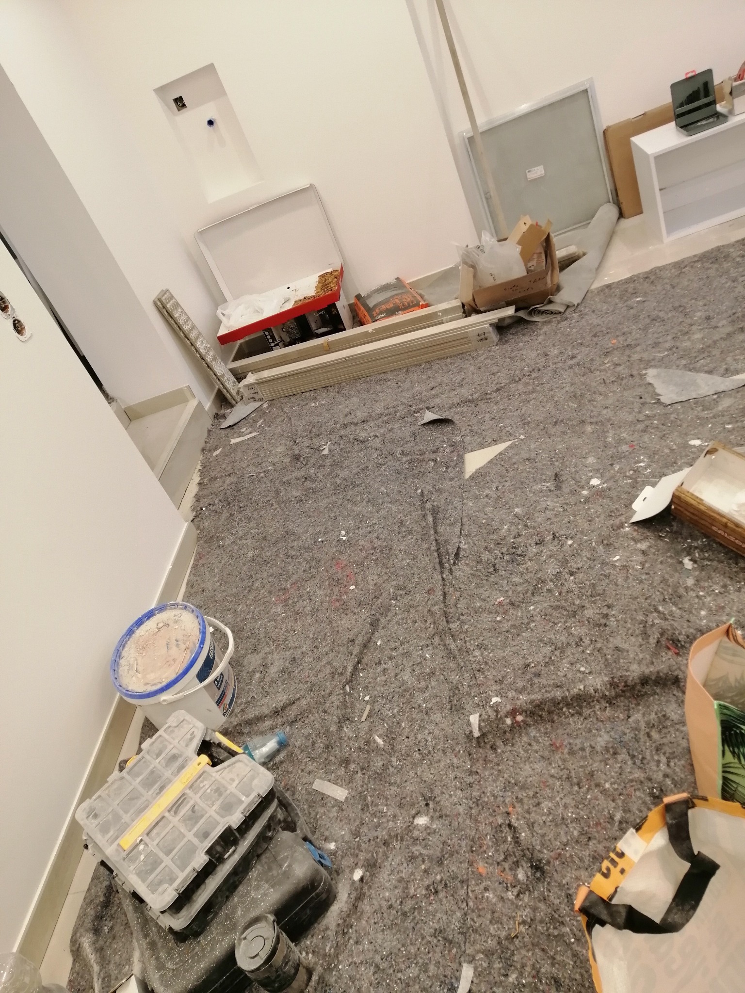 Przygotowanie do remontu: podłoga zabezpieczona szarą folią malarską, widoczne narzędzia, wiadro z farbą, listwy przypodłogowe i materiały budowlane w pomieszczeniu z białymi ścianami.