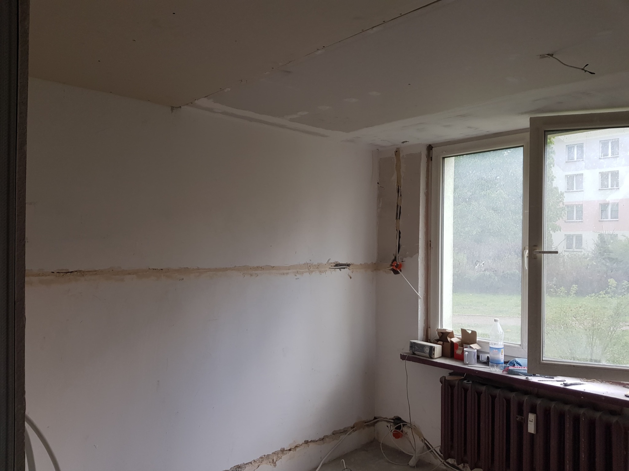 Pokój w trakcie remontu: ściany z widocznymi śladami po instalacjach, częściowo wykończona sufitowa płyta karton-gips, grzejnik pod oknem, narzędzia i materiały budowlane na parapecie.