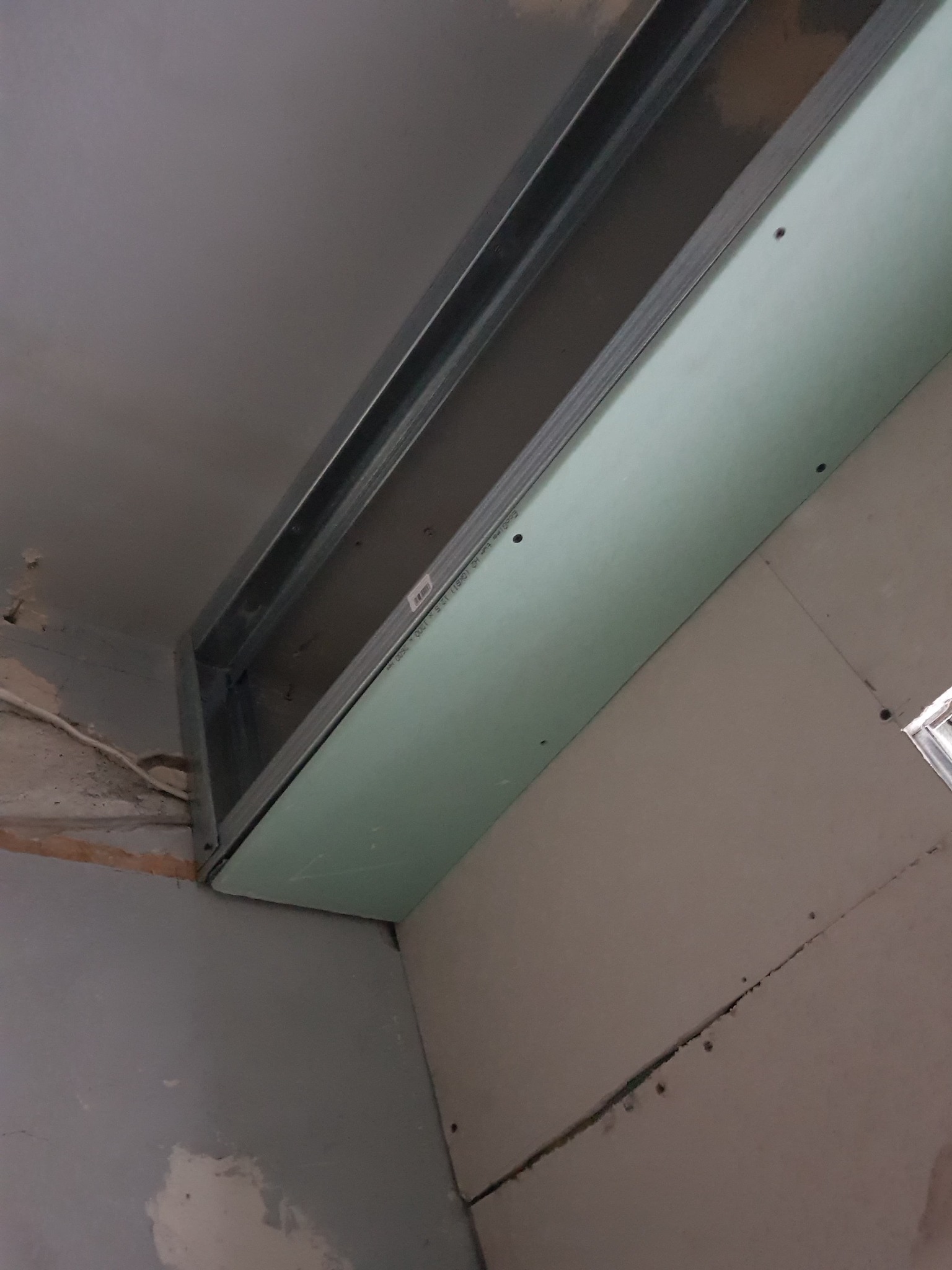 Montaż płyty kartonowo-gipsowej na metalowej konstrukcji sufitowej w trakcie remontu, widoczne śruby mocujące i fragment starej ściany w tle.