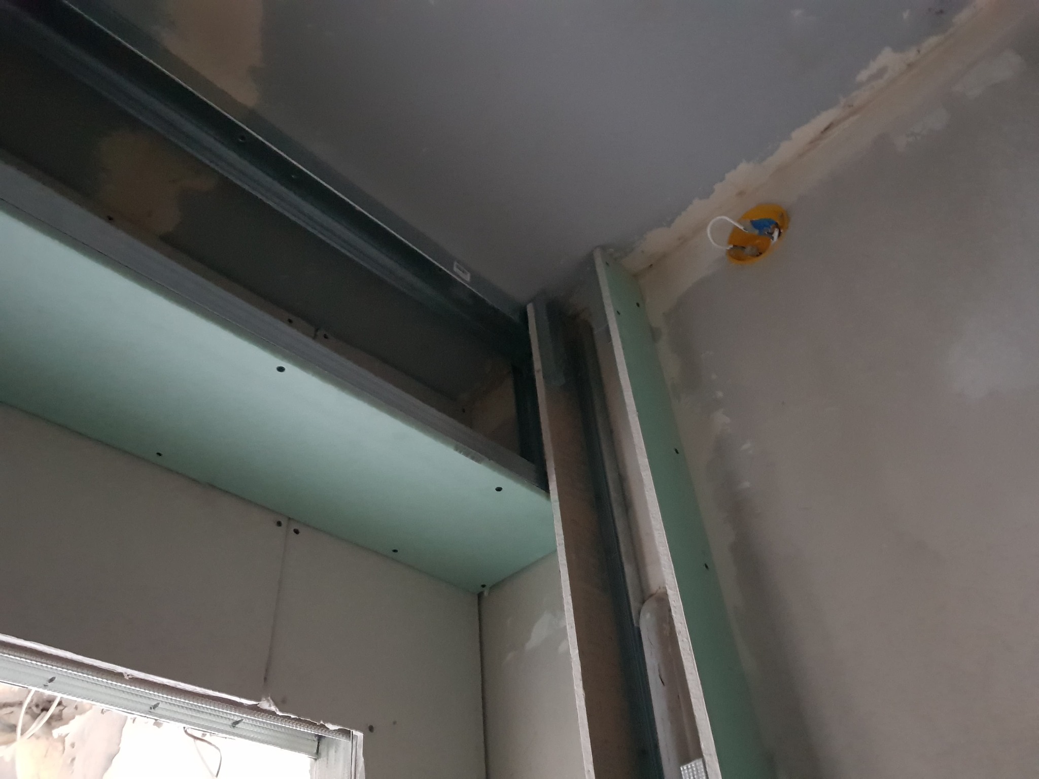 Montaż płyty karton gips na metalowym stelażu, widoczne łączenia płyt i żółta puszka elektryczna w suficie.