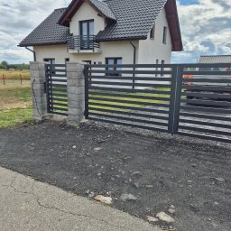 DK STAL Daniel Kaleta - Nowoczesna, pozioma brama wjazdowa z szarymi słupkami z bloczków betonowych, na tle domu z balkonem i pochmurnego nieba.