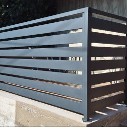 DK STAL Daniel Kaleta - Nowoczesna, pozioma osłona śmietnika z metalowych profili w kolorze grafitowym, zamontowana na betonowym podeście przed domem z drewnianym wykończeniem.