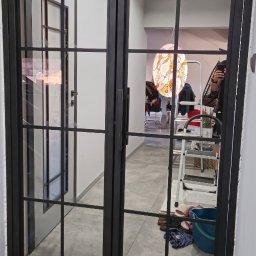 DK STAL Daniel Kaleta - Dwuskrzydłowe drzwi metalowe z czarnymi szprosami, widok na wnętrze z lampą witrażową i rozstawionym sprzętem, niedokończone prace wykończeniowe.