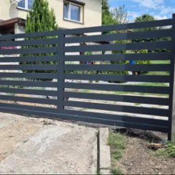 Nowoczesna, przesuwna brama wjazdowa w kolorze antracytowym z poziomymi profilami, zamontowana przy betonowym słupku, widoczna z poziomu gruntu na tle domu jednorodzinnego z ogrodem.