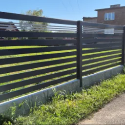 Nowoczesne ogrodzenie posesji z poziomych, szarych paneli metalowych, z białymi akcentami w górnej części, na tle zielonego trawnika i budynków mieszkalnych.