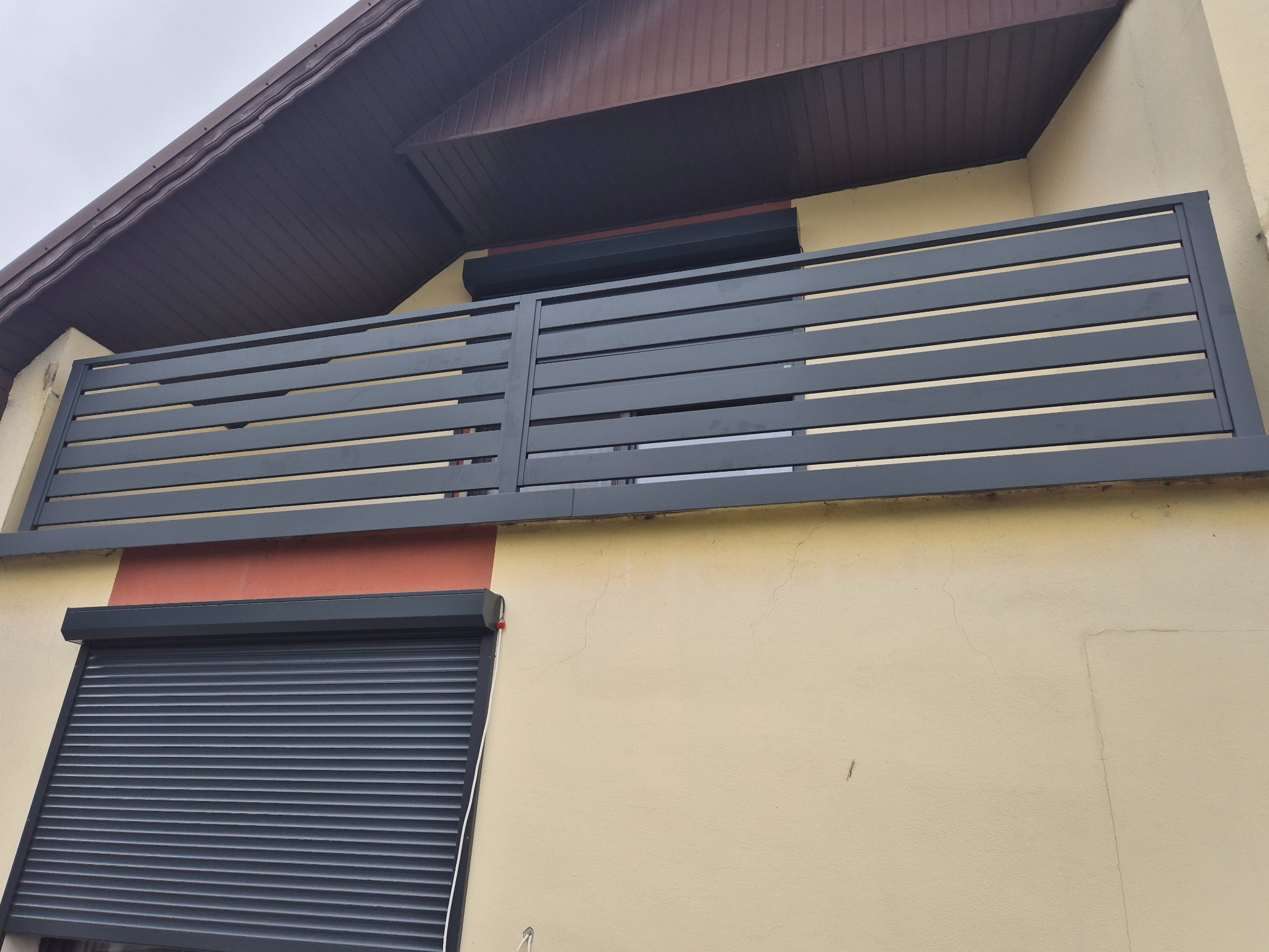 Nowoczesna balustrada balkonowa z poziomymi, ciemnoszarymi profilami, zamontowana na budynku z beżową elewacją i brązowym dachem, widoczna również roleta okienna w tym samym kolorze.