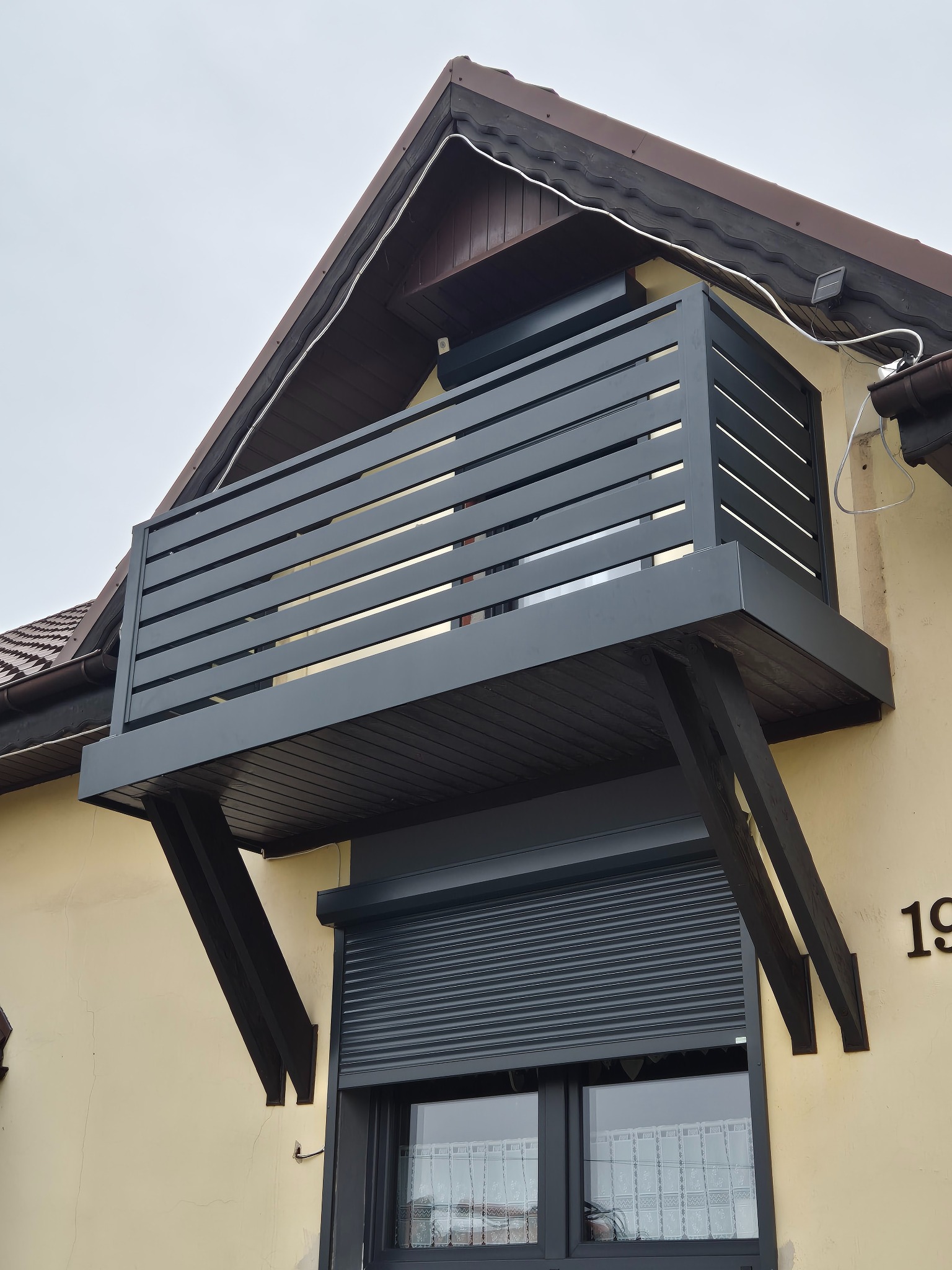 Balkon z szarą, poziomą balustradą z drewnianą podłogą, wsparty na dwóch drewnianych belkach, zamontowany nad oknem z roletą zewnętrzną na ścianie budynku.