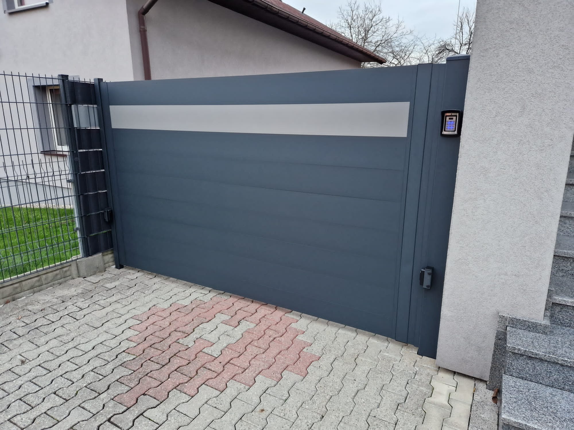 Nowoczesna, przesuwna brama wjazdowa w kolorze grafitowym z poziomym, jasnym pasem, wyposażona w panel sterowania numerycznego, zamontowana na posesji z kostką brukową w odcieniach szarości...