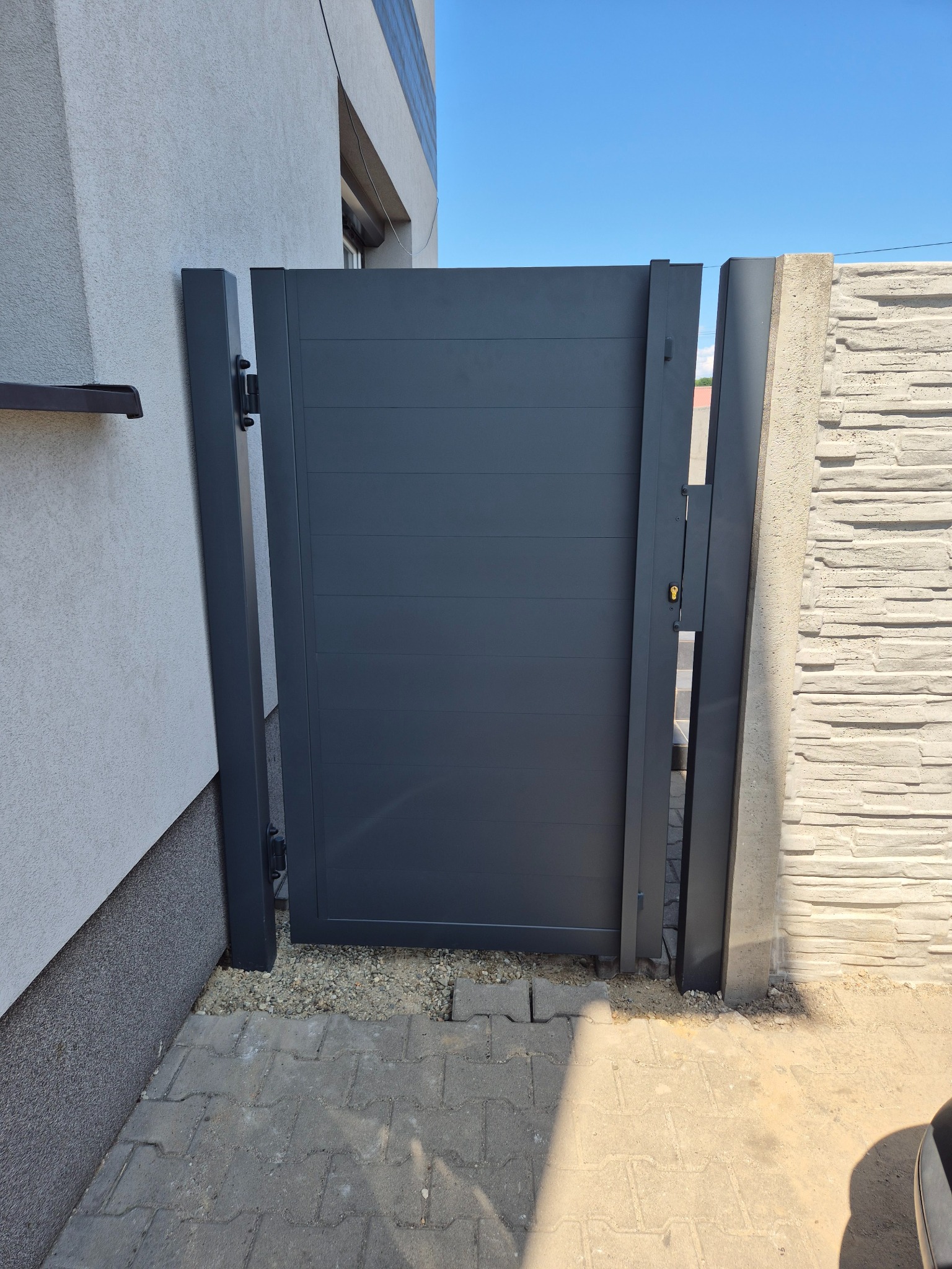 Nowoczesna, pozioma, szara furtka wejściowa z zamkiem, zamontowana między betonowym słupkiem imitującym kamień a ścianą budynku, widok z zewnątrz.