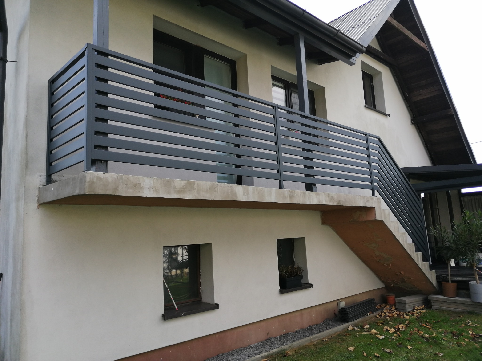 Szara, pozioma balustrada balkonowa z metalowymi szczeblami, widok z dołu na balkon domu z jasną elewacją i schodami z poręczą, jesienne liście na trawniku.