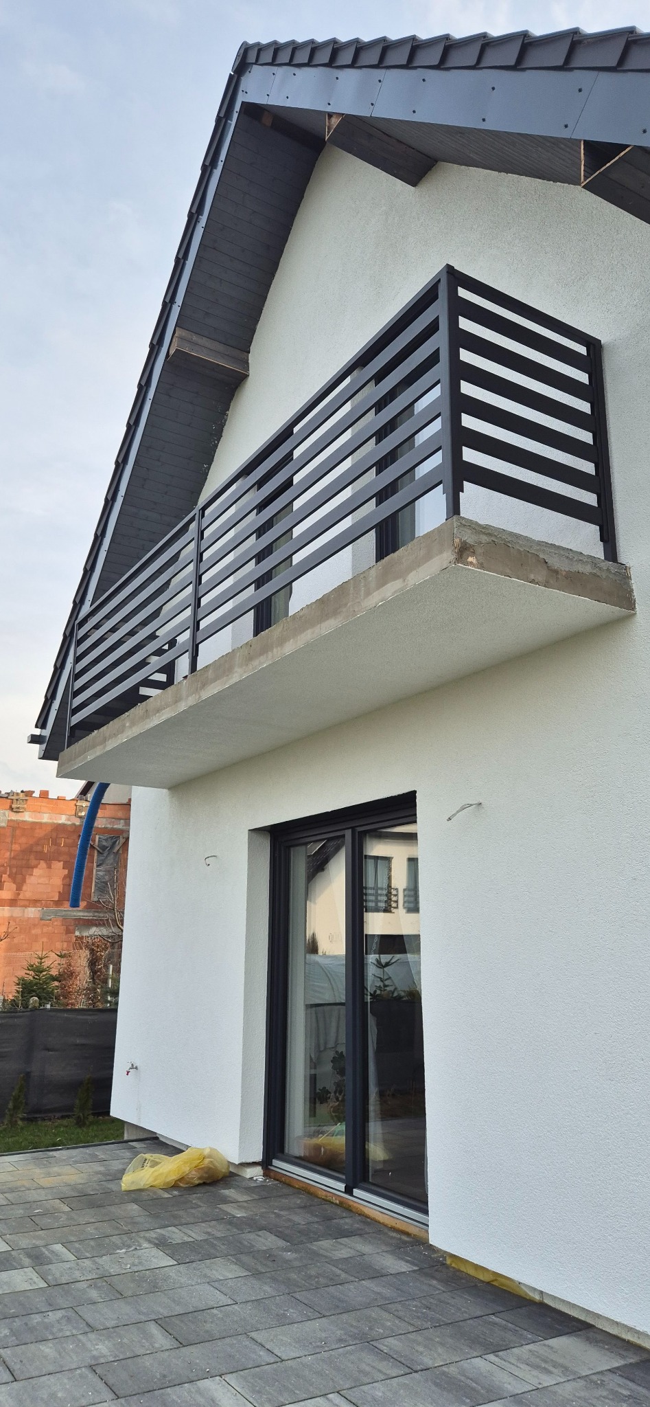Nowoczesny balkon z czarną, poziomą balustradą na tle białej ściany domu z widoczną konstrukcją dachu i fragmentem kostki brukowej.