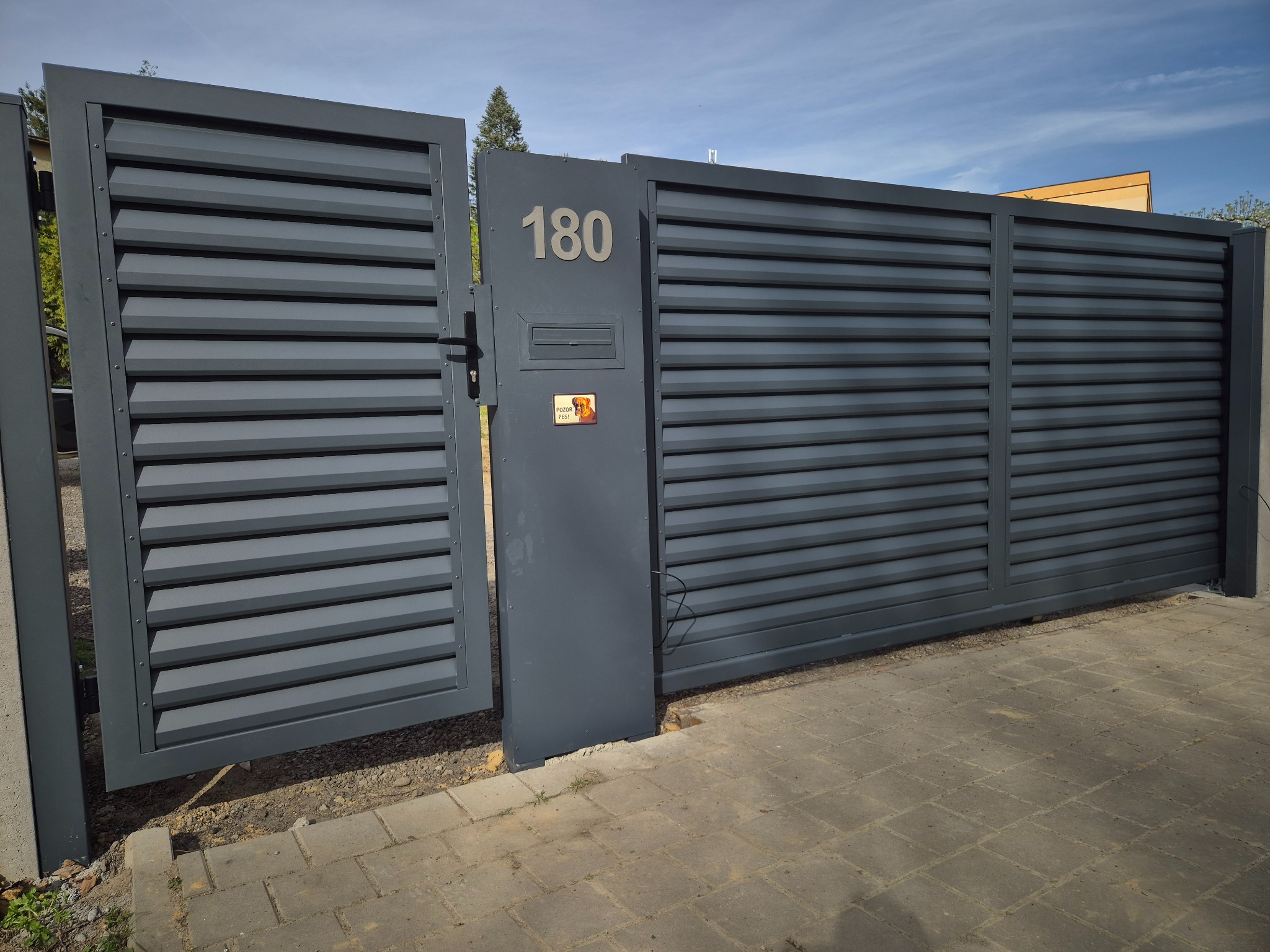 Nowoczesna, przesuwna brama wjazdowa w kolorze grafitowym z poziomymi lamelami, numer domu 180 i skrzynka na listy, częściowo otwarta, na tle błękitnego nieba.