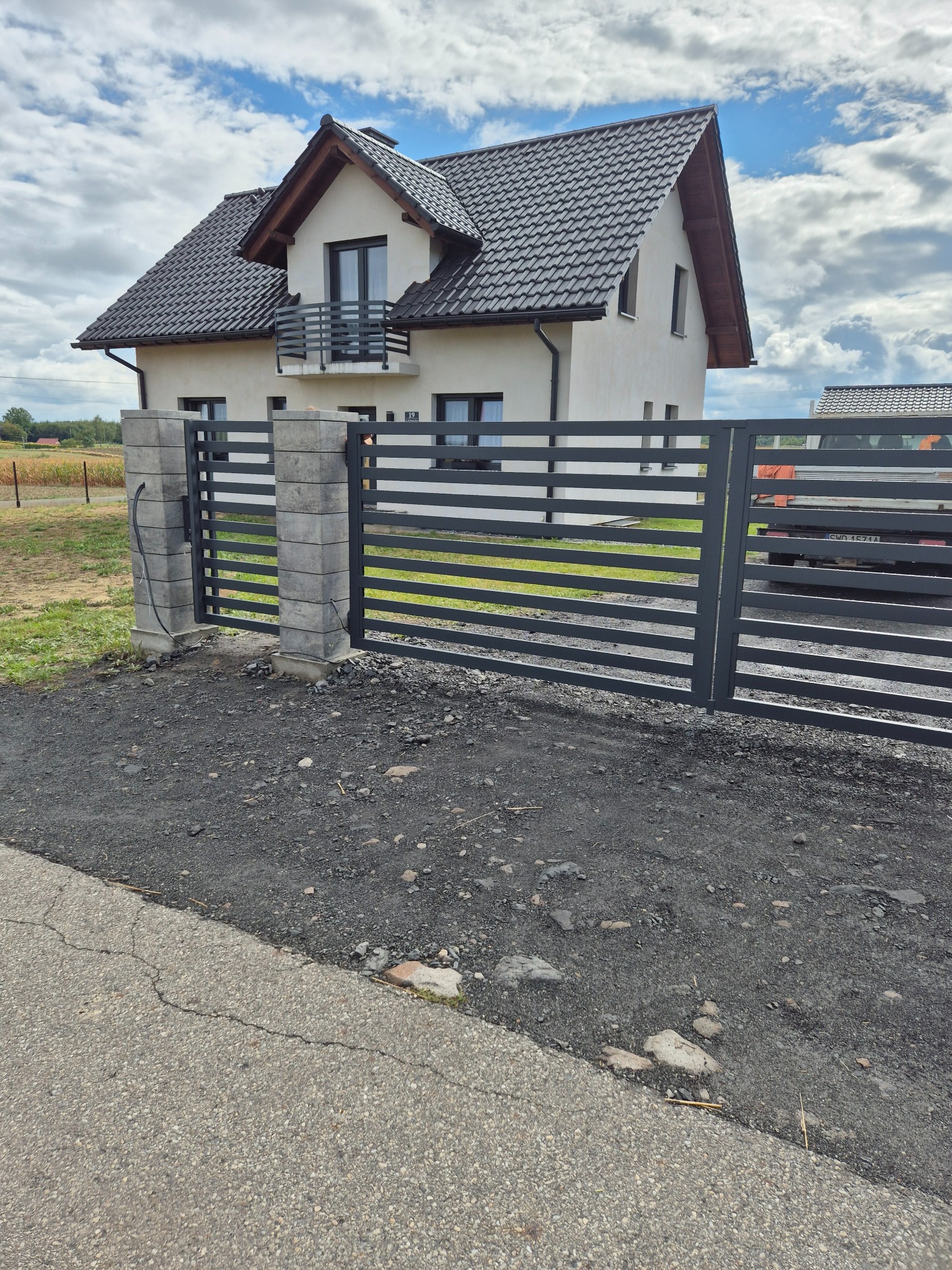 Nowoczesna, pozioma brama wjazdowa z szarymi słupkami z bloczków betonowych, na tle domu z balkonem i pochmurnego nieba.