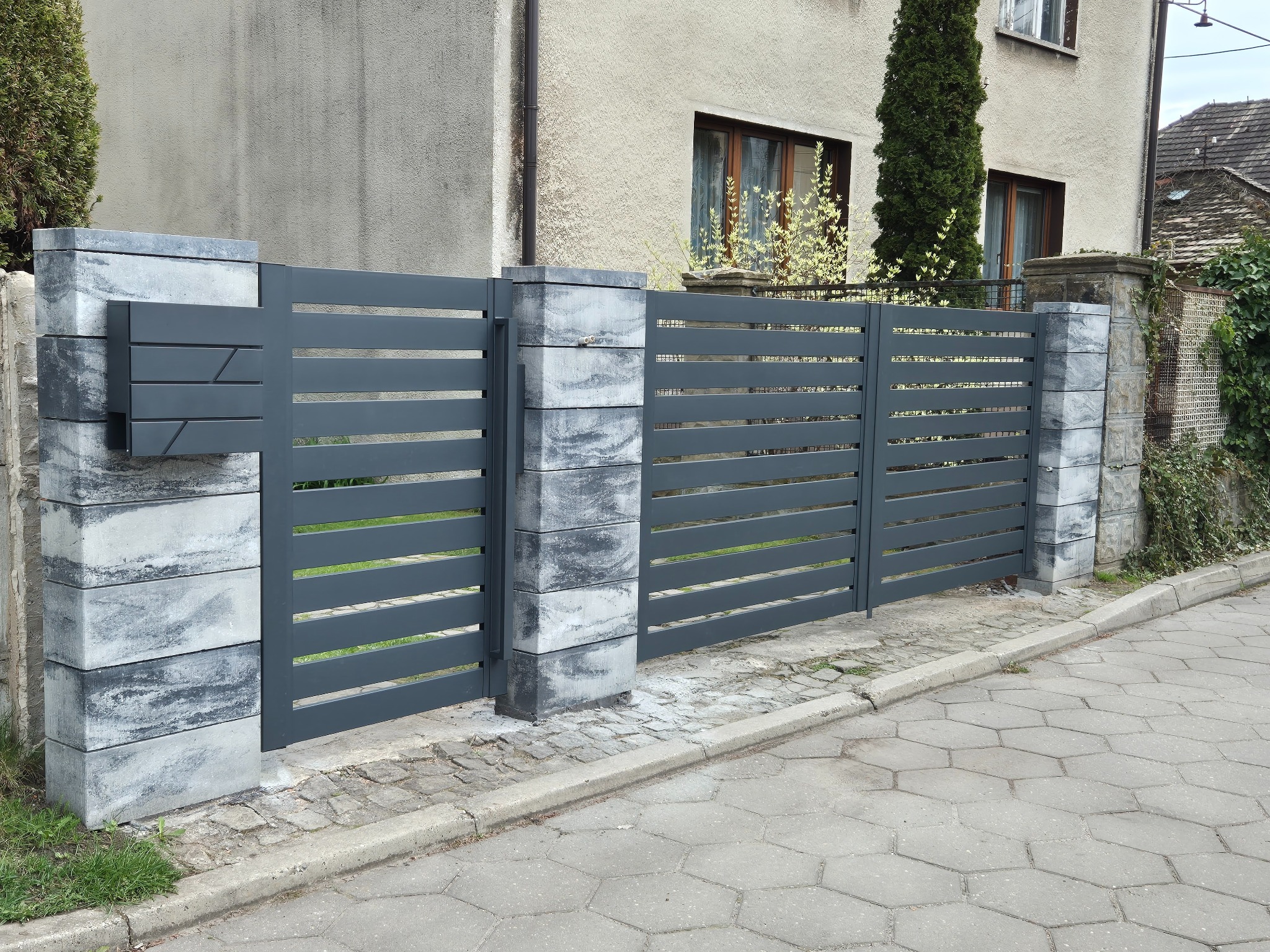Nowoczesna, pozioma brama wjazdowa w kolorze grafitowym z szarymi, betonowymi słupkami, uzupełniona o skrzynkę na listy, na tle domu z ogrodem.