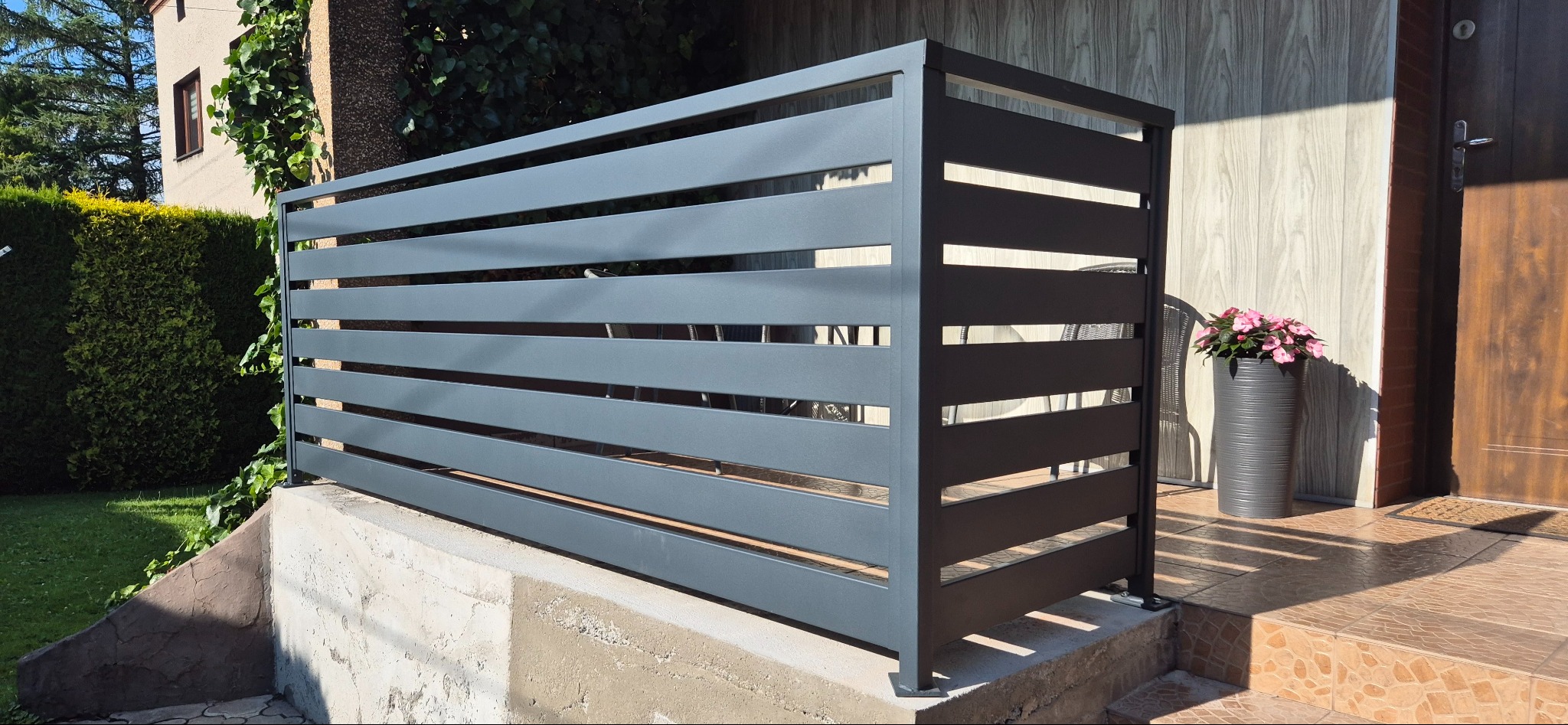 Nowoczesna, pozioma osłona śmietnika z metalowych profili w kolorze grafitowym, zamontowana na betonowym podeście przed domem z drewnianym wykończeniem.