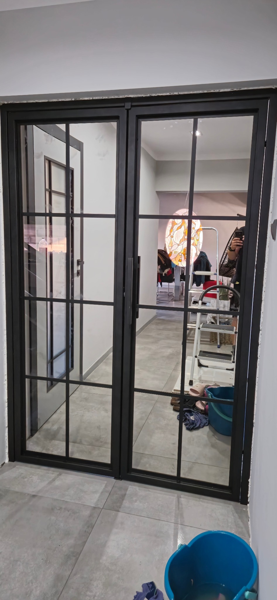 Dwuskrzydłowe drzwi metalowe z czarnymi szprosami, widok na wnętrze z lampą witrażową i rozstawionym sprzętem, niedokończone prace wykończeniowe.