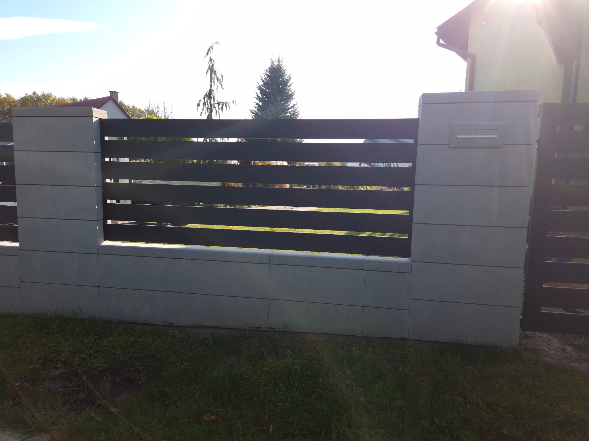 Nowoczesne ogrodzenie panelowe z elementami betonowymi i metalowymi, widok z zewnątrz na tle trawnika i zabudowań.