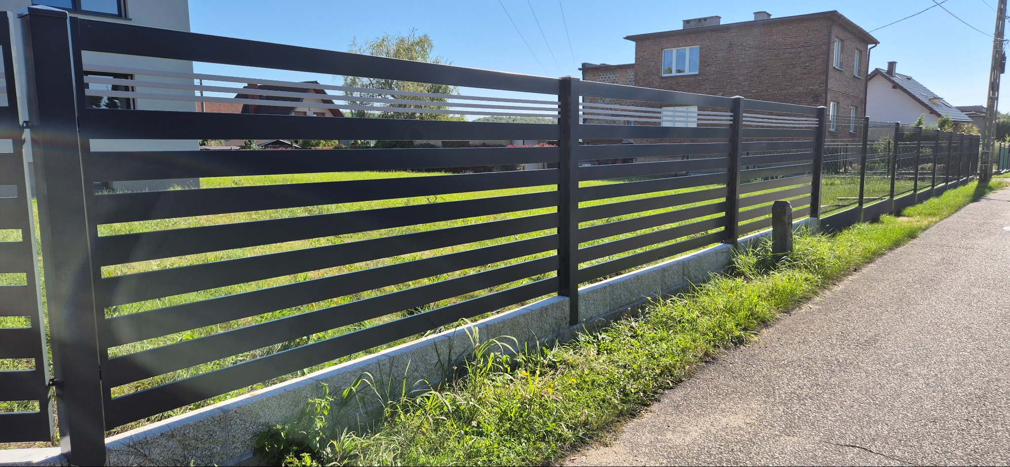 Nowoczesne ogrodzenie posesji z poziomych, szarych paneli metalowych, z białymi akcentami w górnej części, na tle zielonego trawnika i budynków mieszkalnych.
