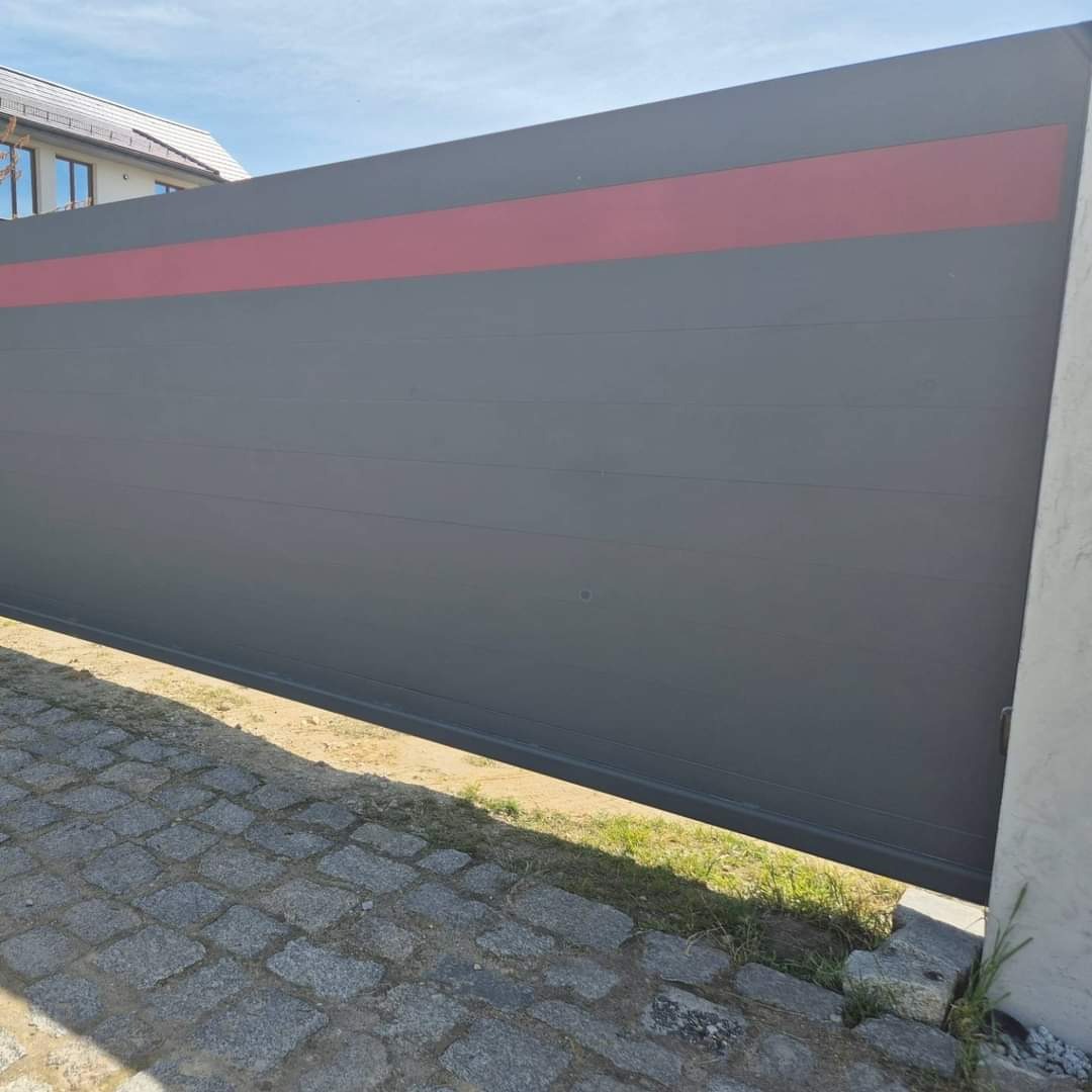Nowoczesna, przesuwna brama w kolorze grafitowym z poziomym czerwonym pasem, widoczna z perspektywy chodnika z kostki brukowej i fragmentu trawnika, z domem w tle.