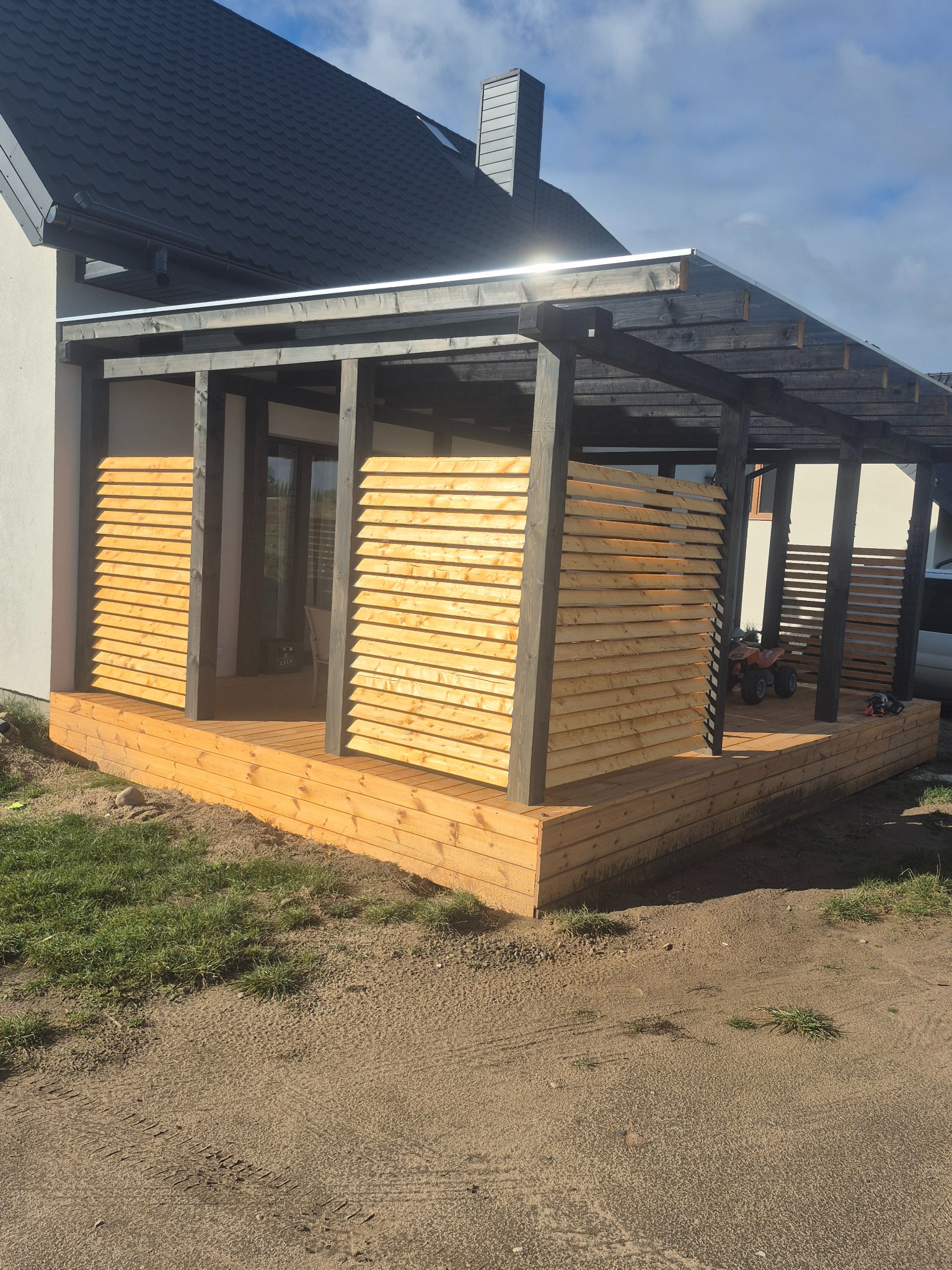 Drewniany taras z zadaszeniem i ażurowymi ściankami, świeżo po budowie, z widocznym quadem dla dziecka. Ujęcie z poziomu gruntu, podkreślające solidność konstrukcji.